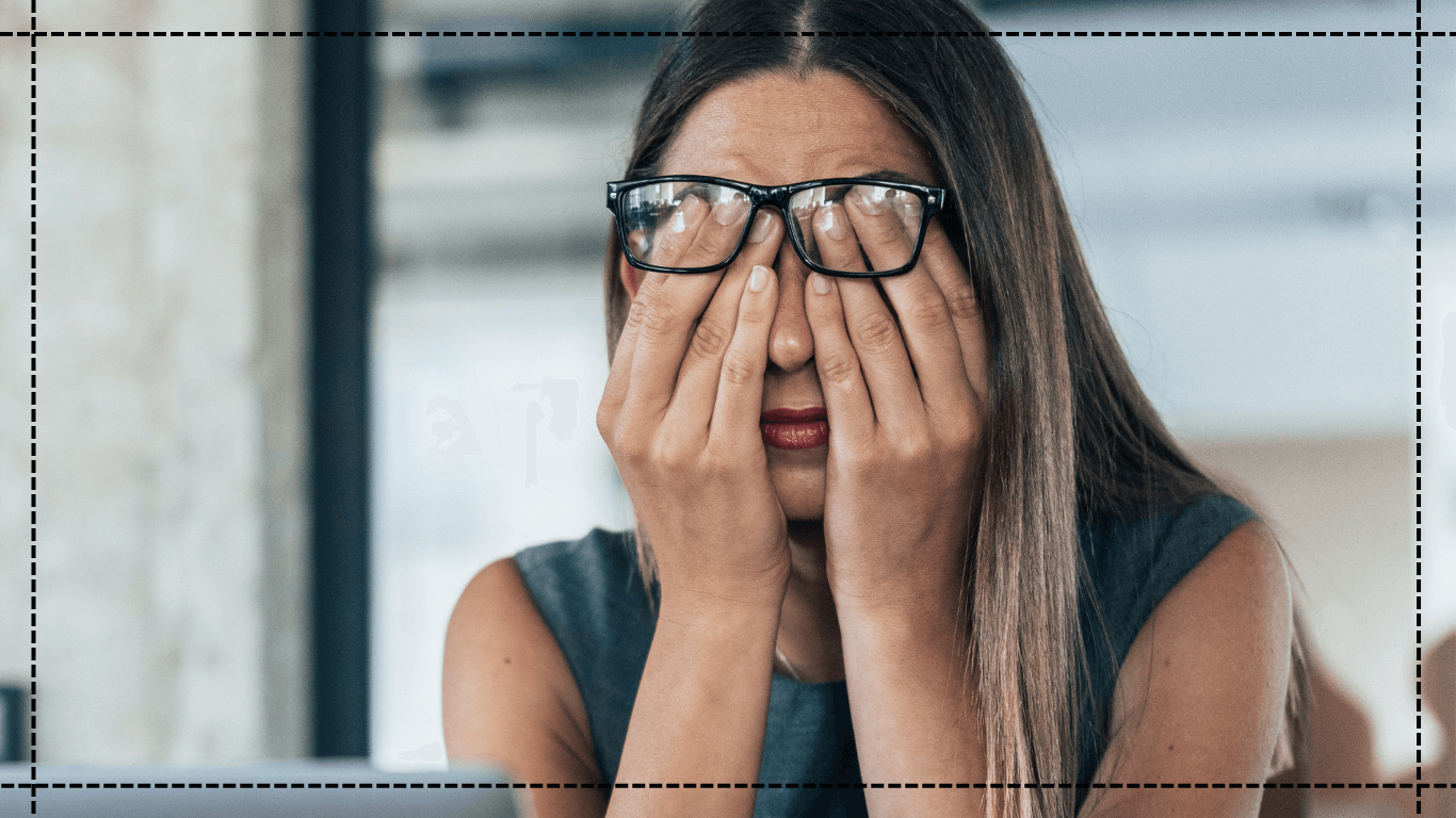 Woman wearing glasses covering her face with her hands in a blurred indoor setting