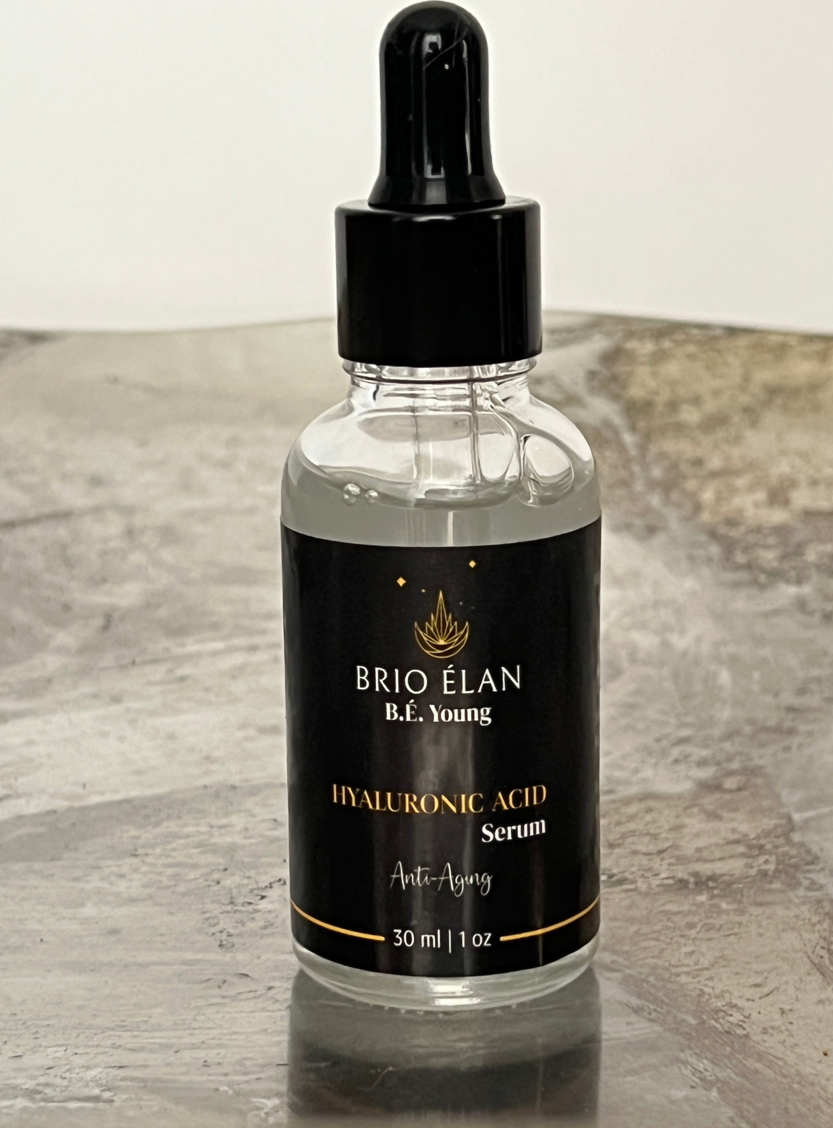 A 30 mL clear dropper bottle of BRIO ÉLAN Hyaluronic Acid Serum presented on a tan marble table top. 